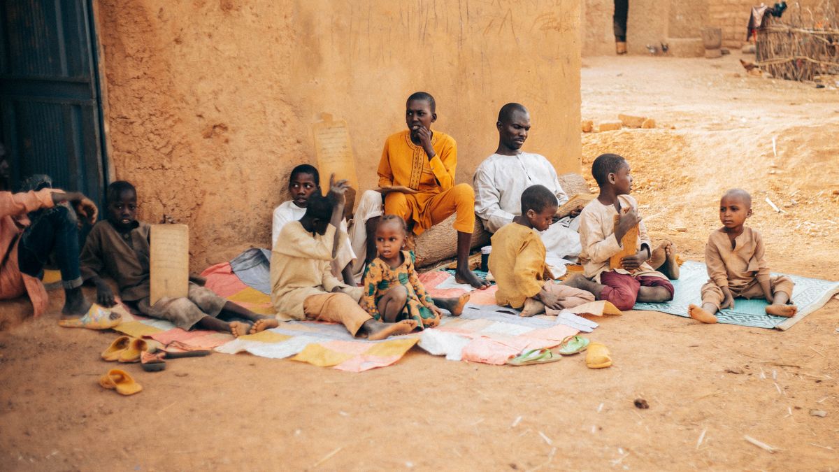 A group of African children and elders engaged in learning activities in a rural outdoor setting.
