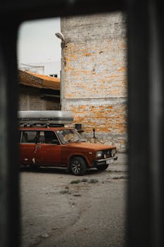 A vintage red car parked beside a weathered brick wall in an urban setting.