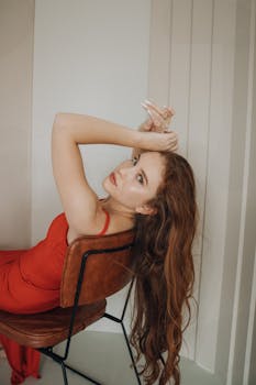 Stylish woman with long hair in red dress posing elegantly indoors on a chair.