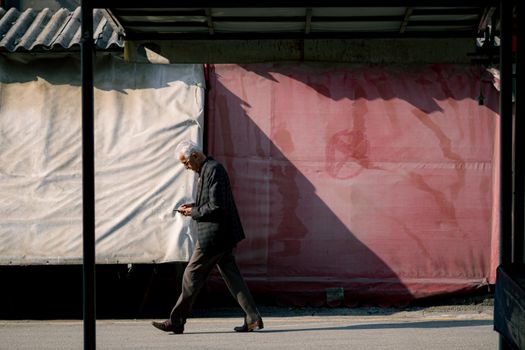 An elderly man walks thoughtfully through a sunlit urban alley, casting a long shadow.