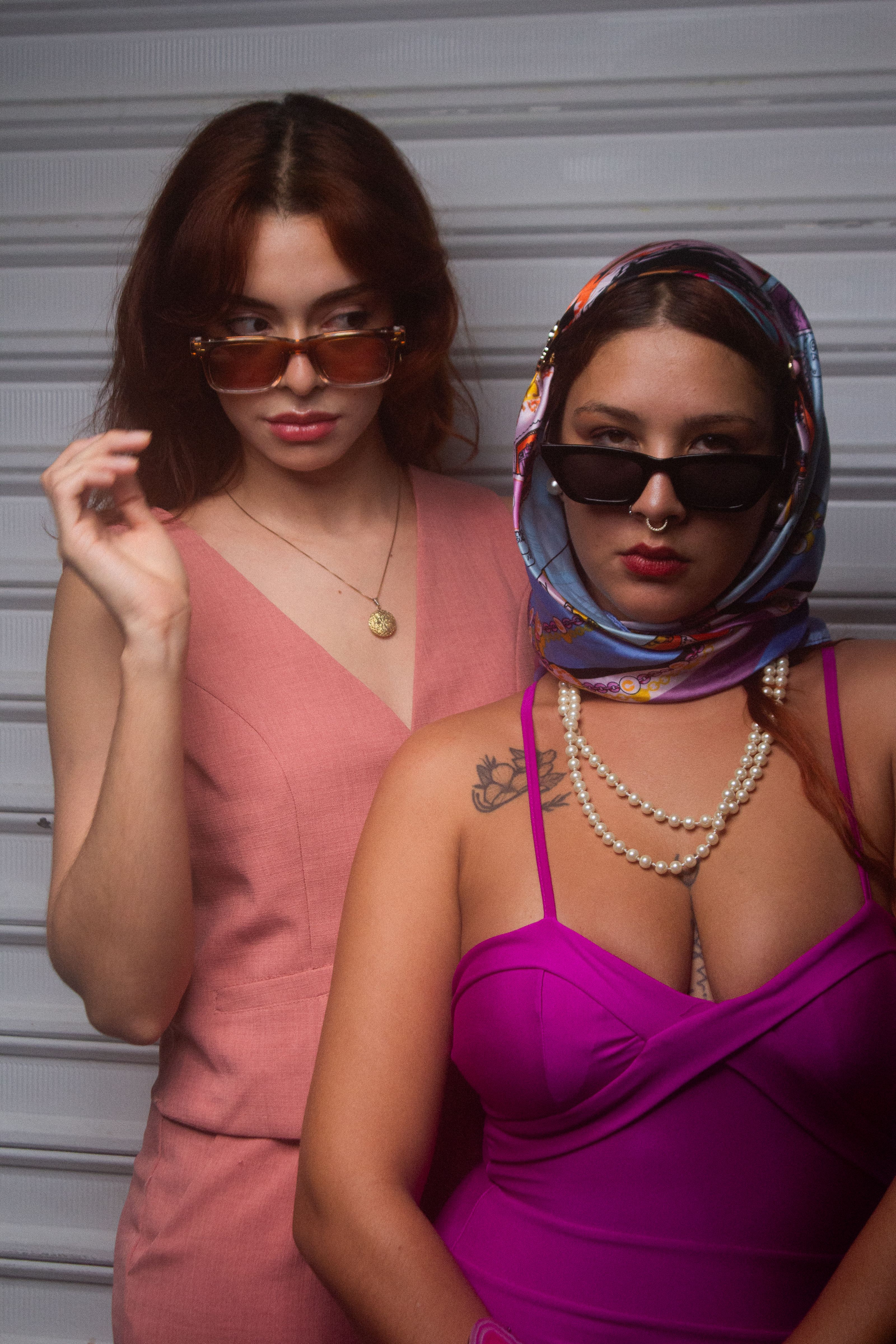 Duas mulheres em trajes elegantes e óculos de sol posando com confiança dentro de um estúdio.