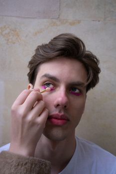 Close-up portrait of a man applying vibrant pink eye makeup with a brush indoors.
