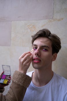 A young man having colorful makeup applied indoors, showcasing beauty care.