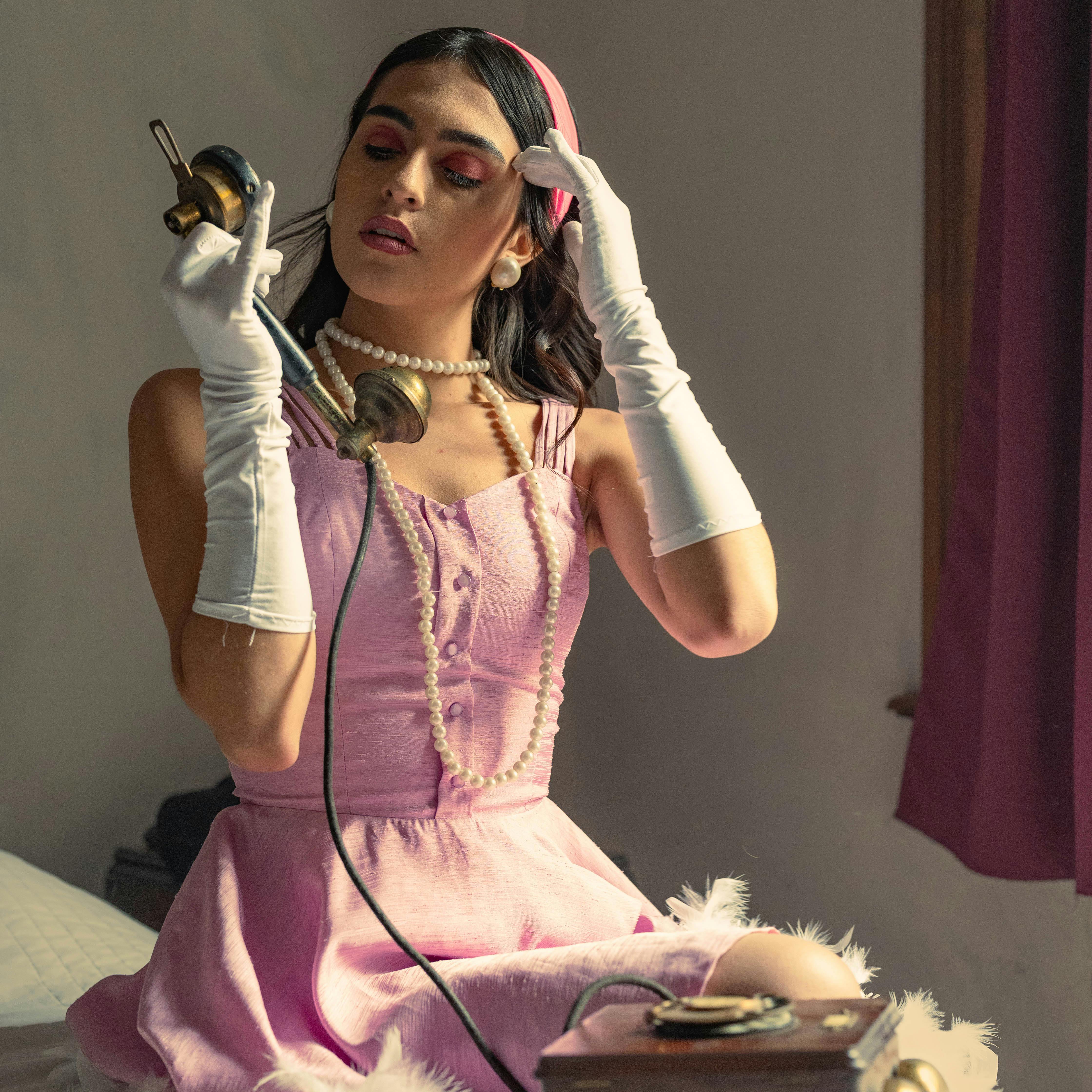 Elegant woman in pink vintage attire posing with a retro telephone indoors.