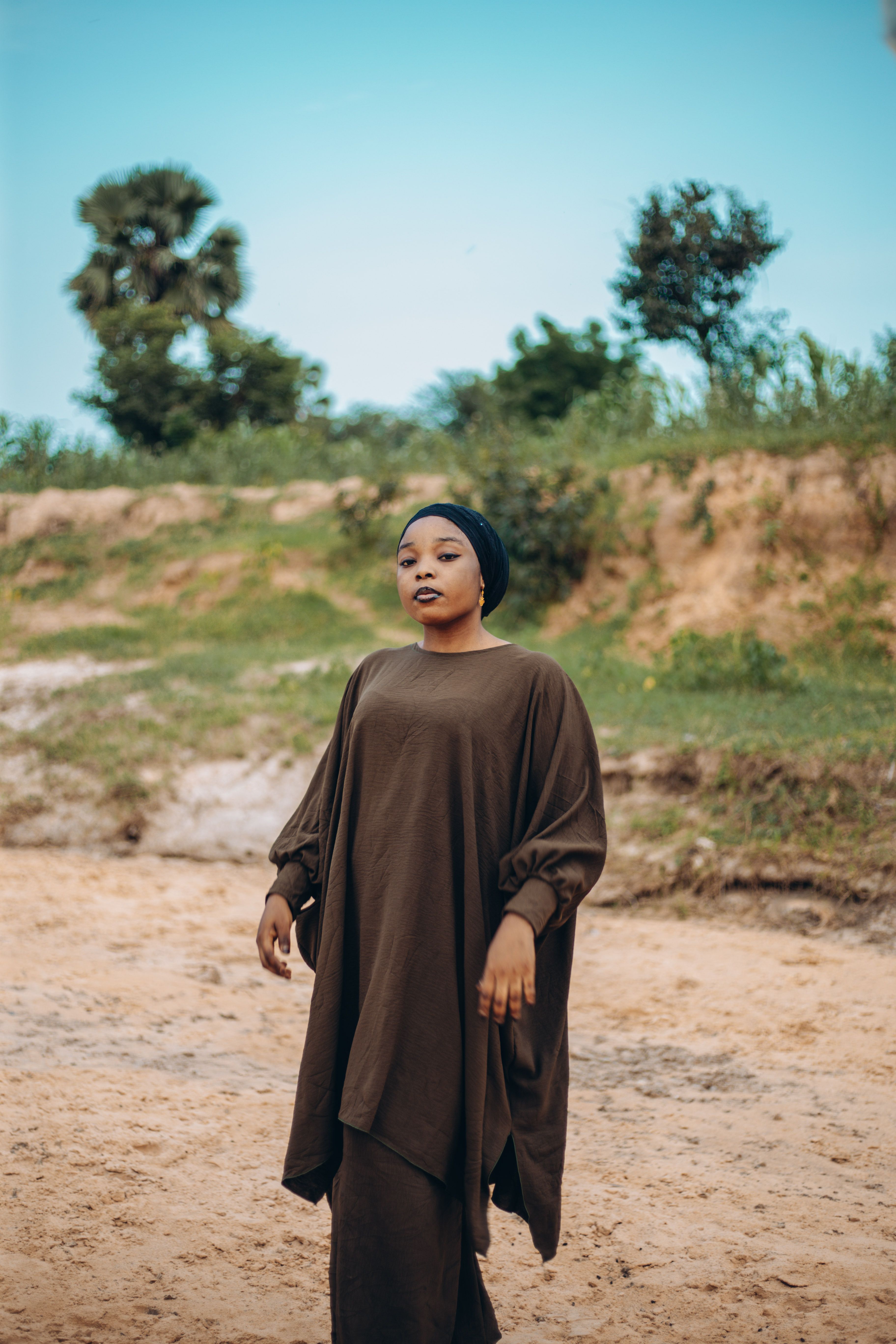 A woman stands gracefully in a brown traditional outfit outdoors, exuding elegance.