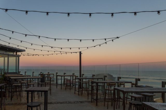 A rooftop terrace strung with festoon lights, tables set for the evening above the ocean at dusk