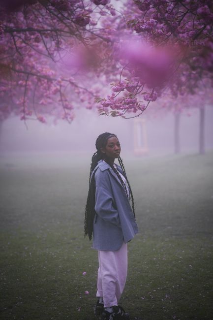 A woman in a stylish coat stands under cherry blossoms in a foggy Berlin park.