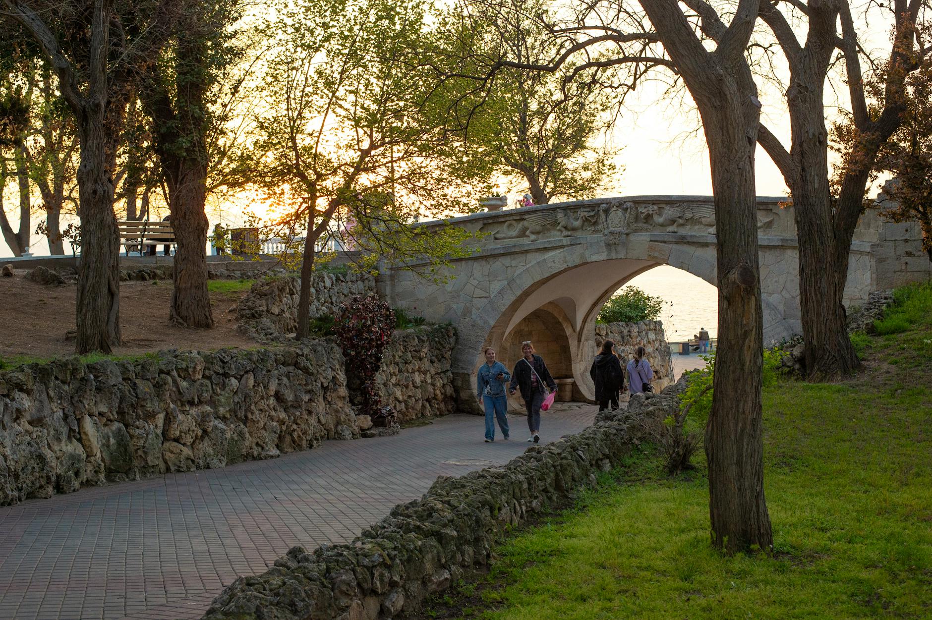 People enjoying a stroll under a picturesque stone bridge in a park at sunset with lush greenery.