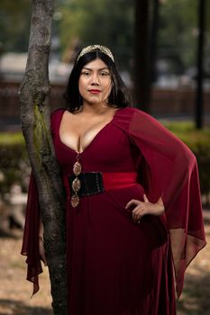 A woman in a red dress poses confidently next to a tree in a park setting.