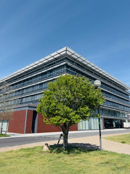 Modern glass office building with green lawn and tree under a clear blue sky.