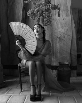 Elegant black and white portrait of a woman seated indoors with a hand fan, exuding timeless grace.