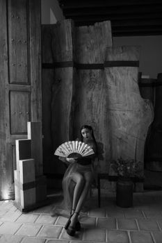 A woman gracefully sits with a fan in a vintage room, exuding elegance and timeless beauty.