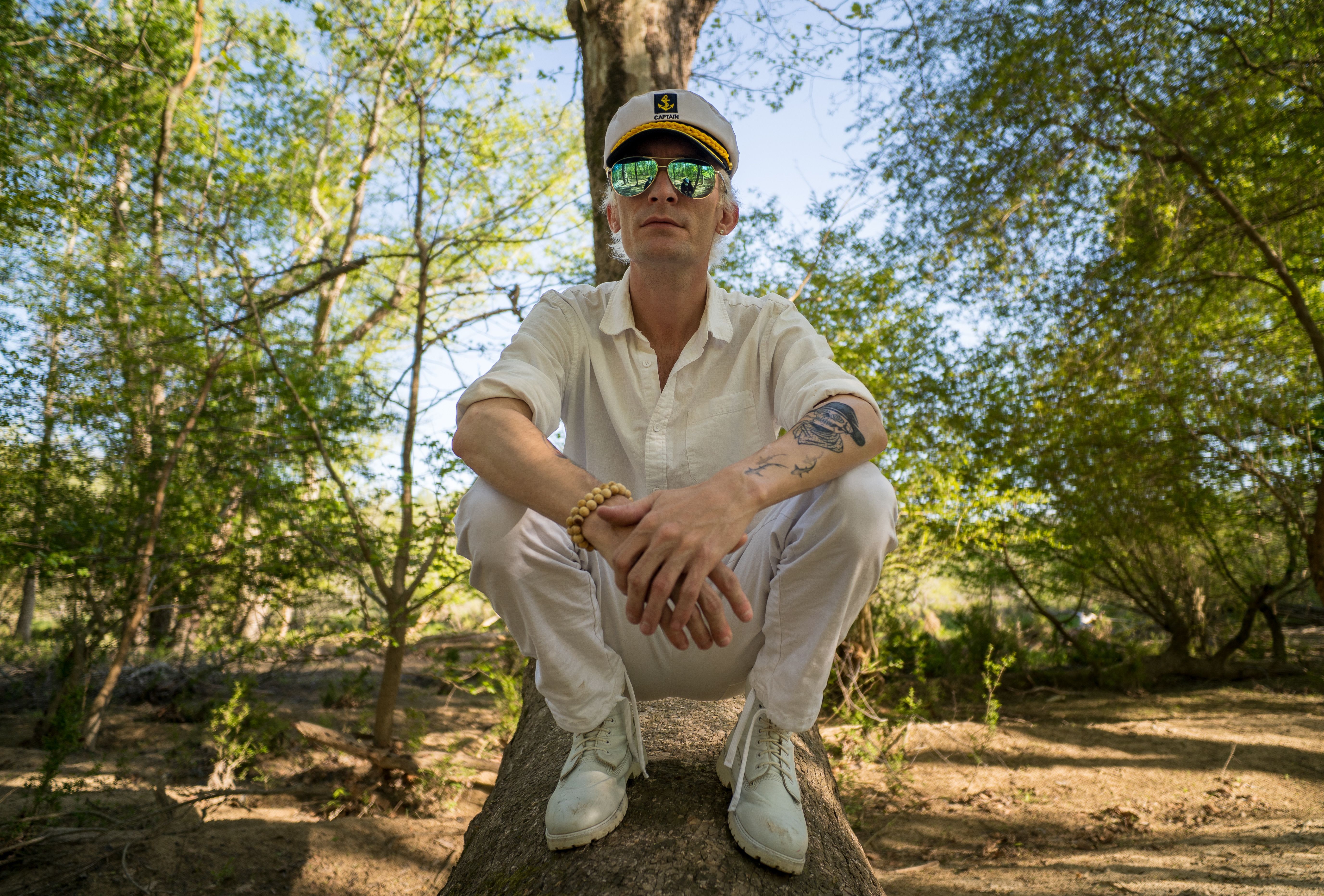 Man in white outfit with captain hat sitting on a tree branch in a lush forest setting.