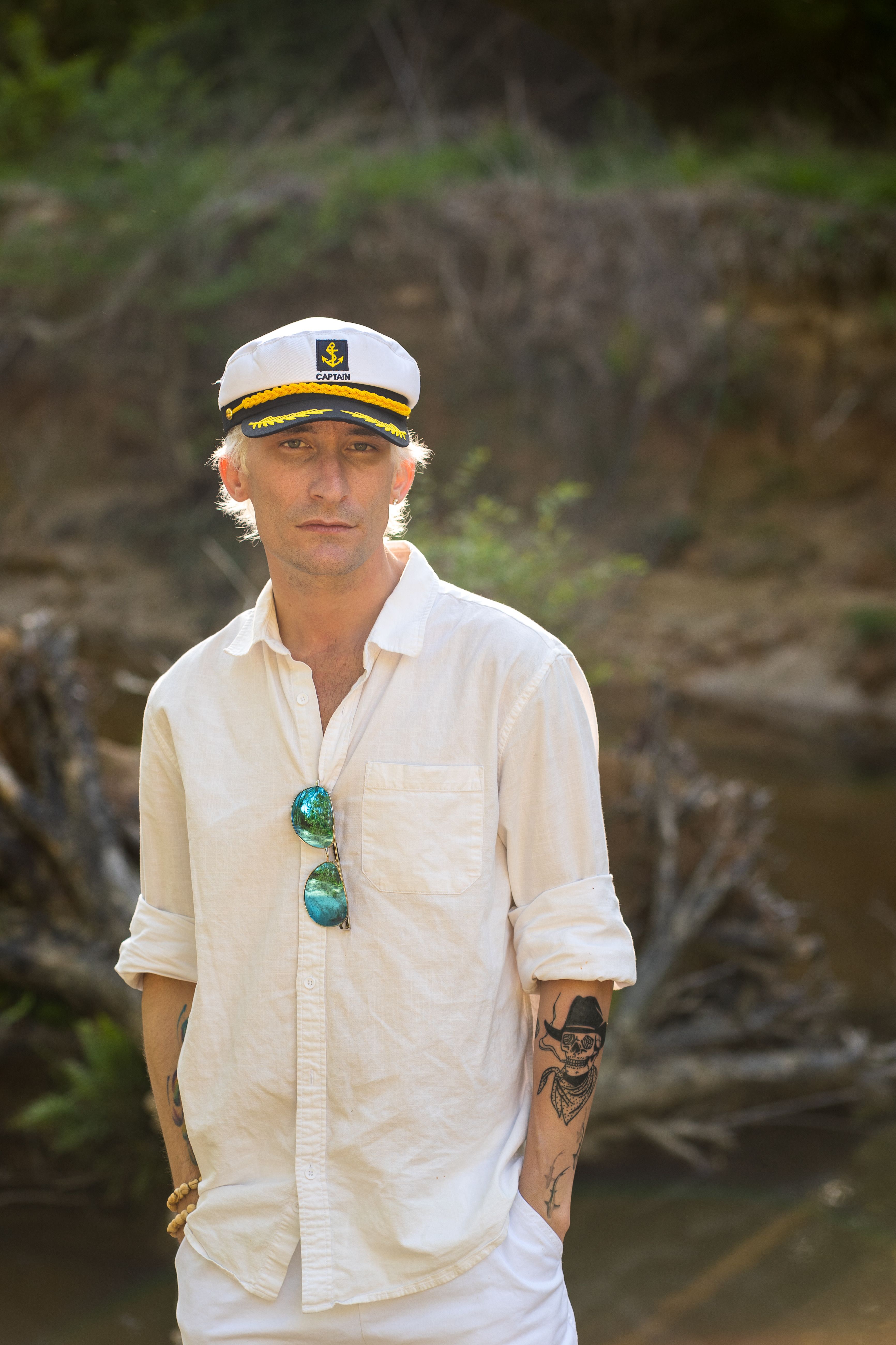 A young man in a captain's hat poses by the riverside, exuding a relaxed nautical style.