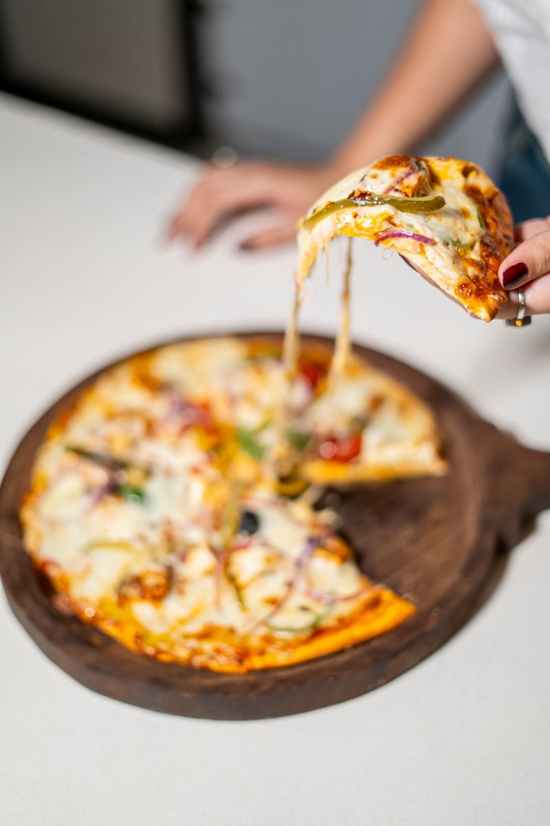 Close-up of a hand taking a slice of cheesy pizza with various toppings on a wooden board.