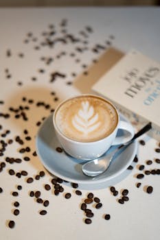A cup of coffee with latte art surrounded by coffee beans and a book in a cozy setup.