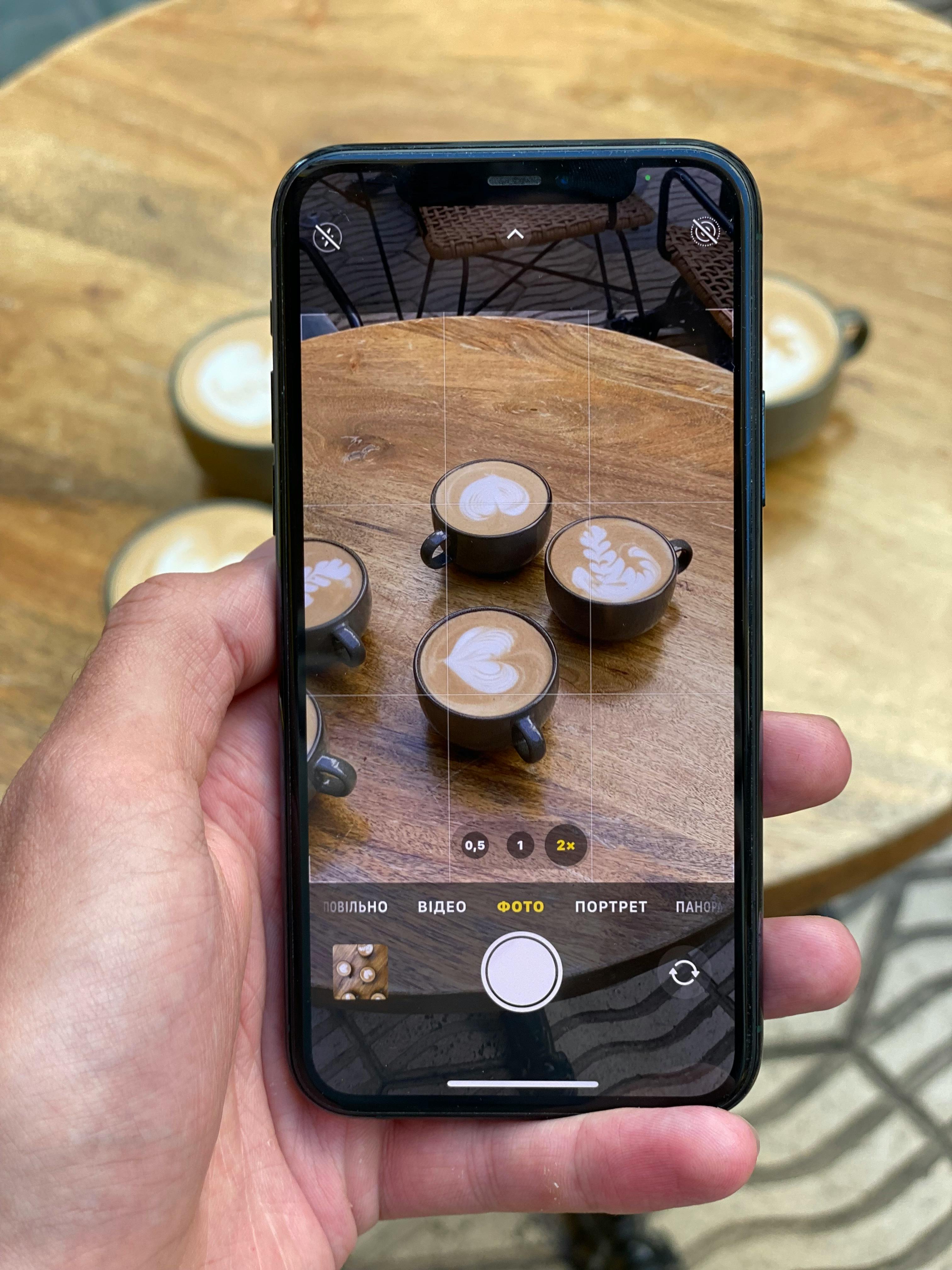 Free A hand holds a smartphone displaying cappuccinos with latte art on a wooden table. Stock Photo