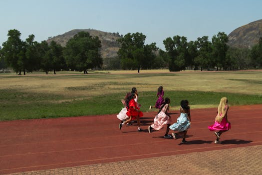 A group of girls wearing colorful dresses running on a track outdoors with hills in the background.
