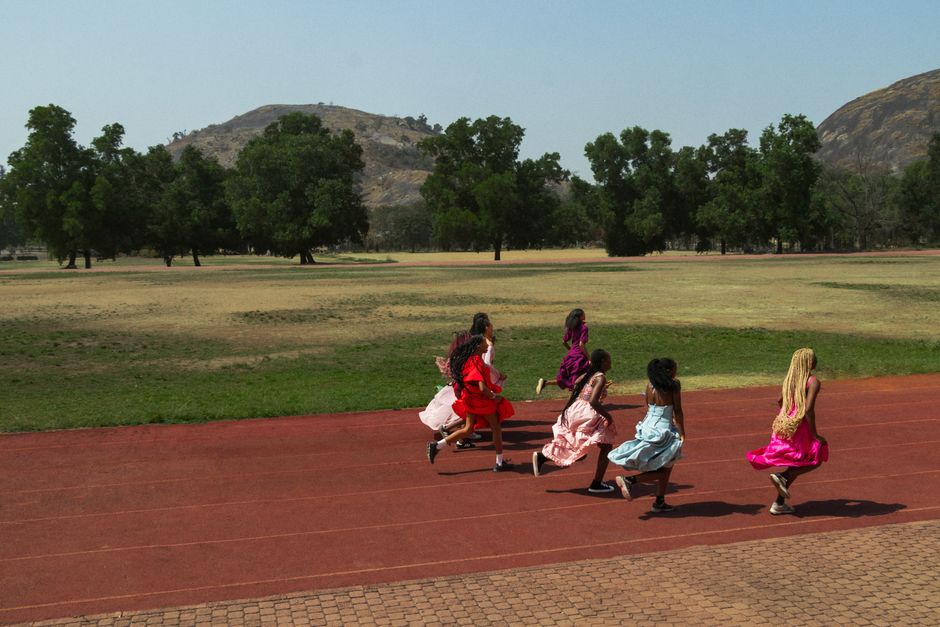 A group of girls wearing colorful dresses running on a track outdoors with hills in the background.