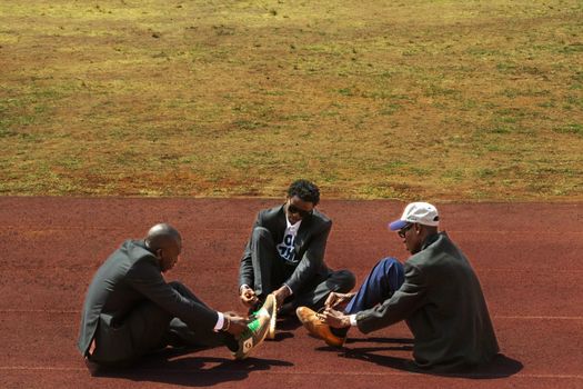 Three men in suits removing their shoes on an outdoor track field.