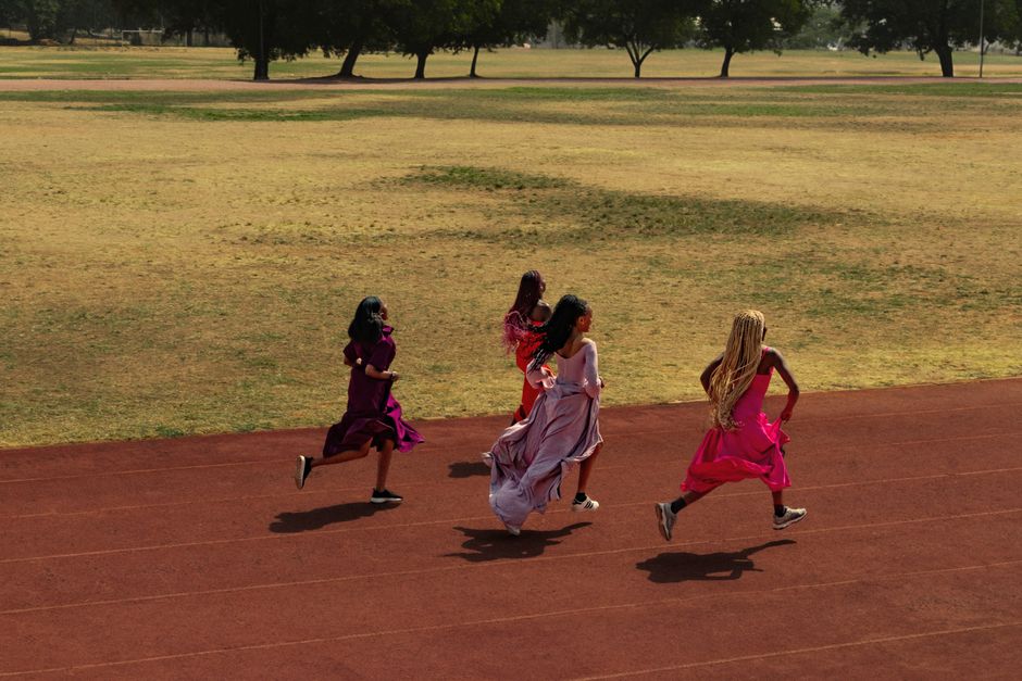 Four women in colorful dresses race on a track in a sunny outdoor setting showcasing motion and energy.