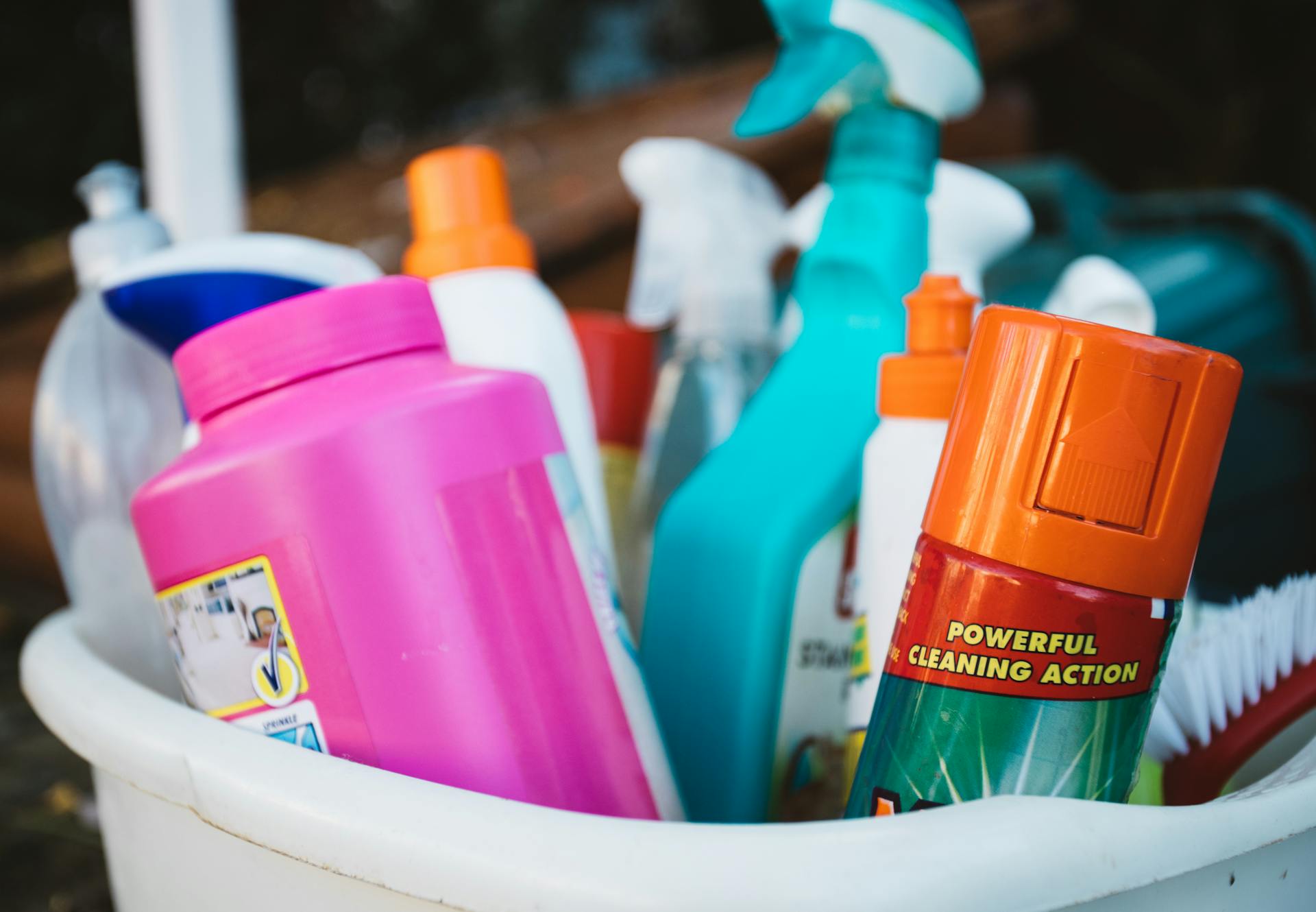 Close-up of assorted cleaning supplies in a bucket outdoors, showcasing various detergents and sprays.