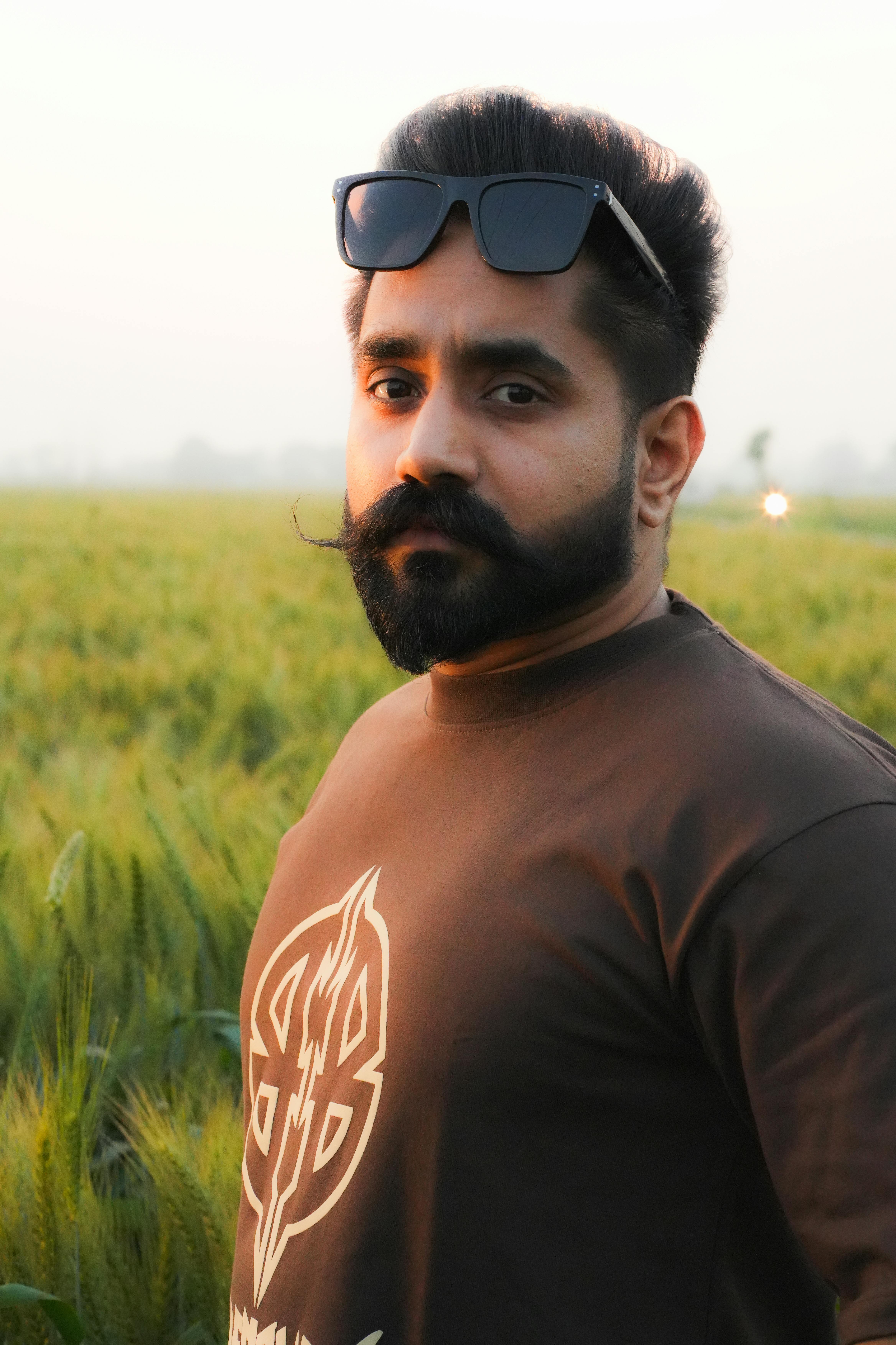 Portrait of a bearded man wearing sunglasses, standing in a lush green wheat field during daylight.