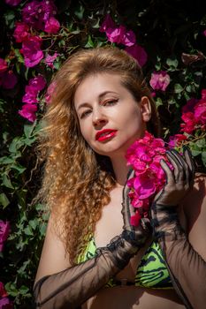 A woman with curly hair and red lips stands among vibrant pink bougainvillea flowers.