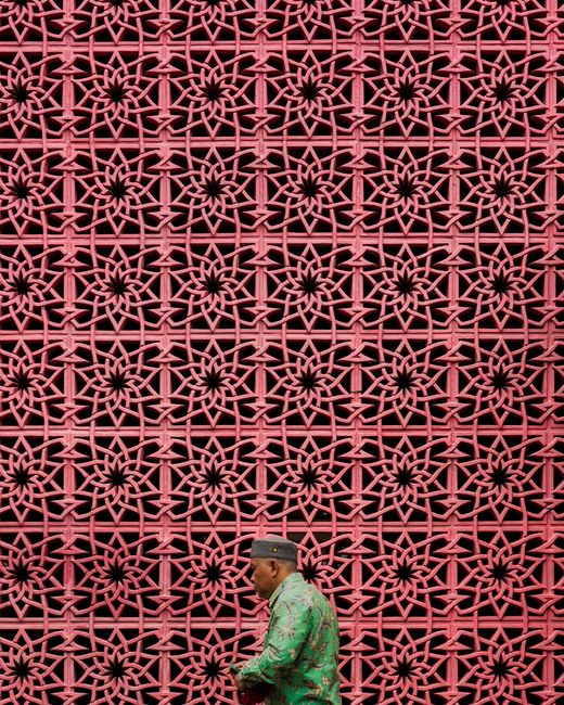 A man walks past a decorative lattice wall in Jakarta, Indonesia, showcasing urban culture and traditional attire.