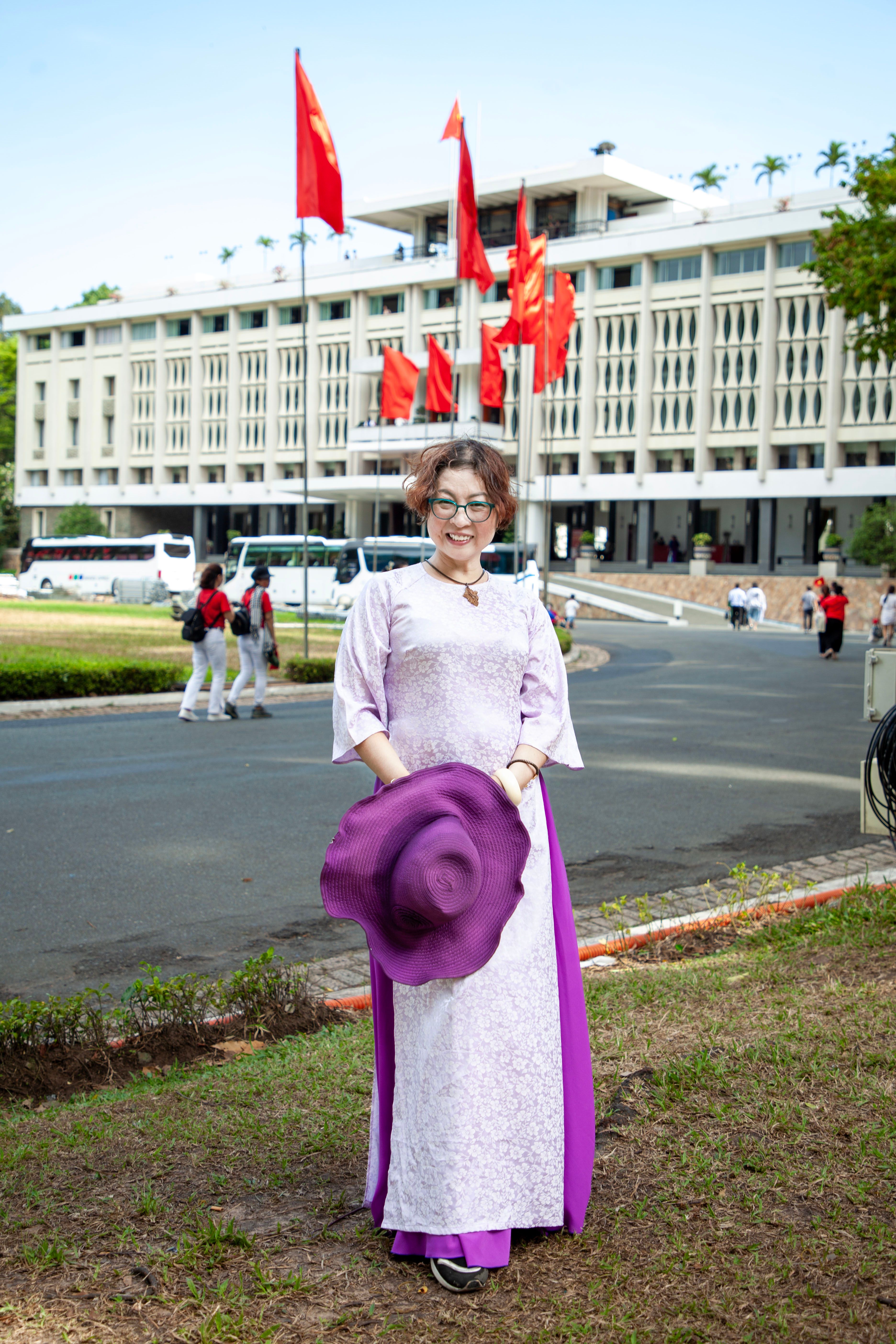 Woman in traditional Ao Dai holds a purple hat outside a government building with red flags.
