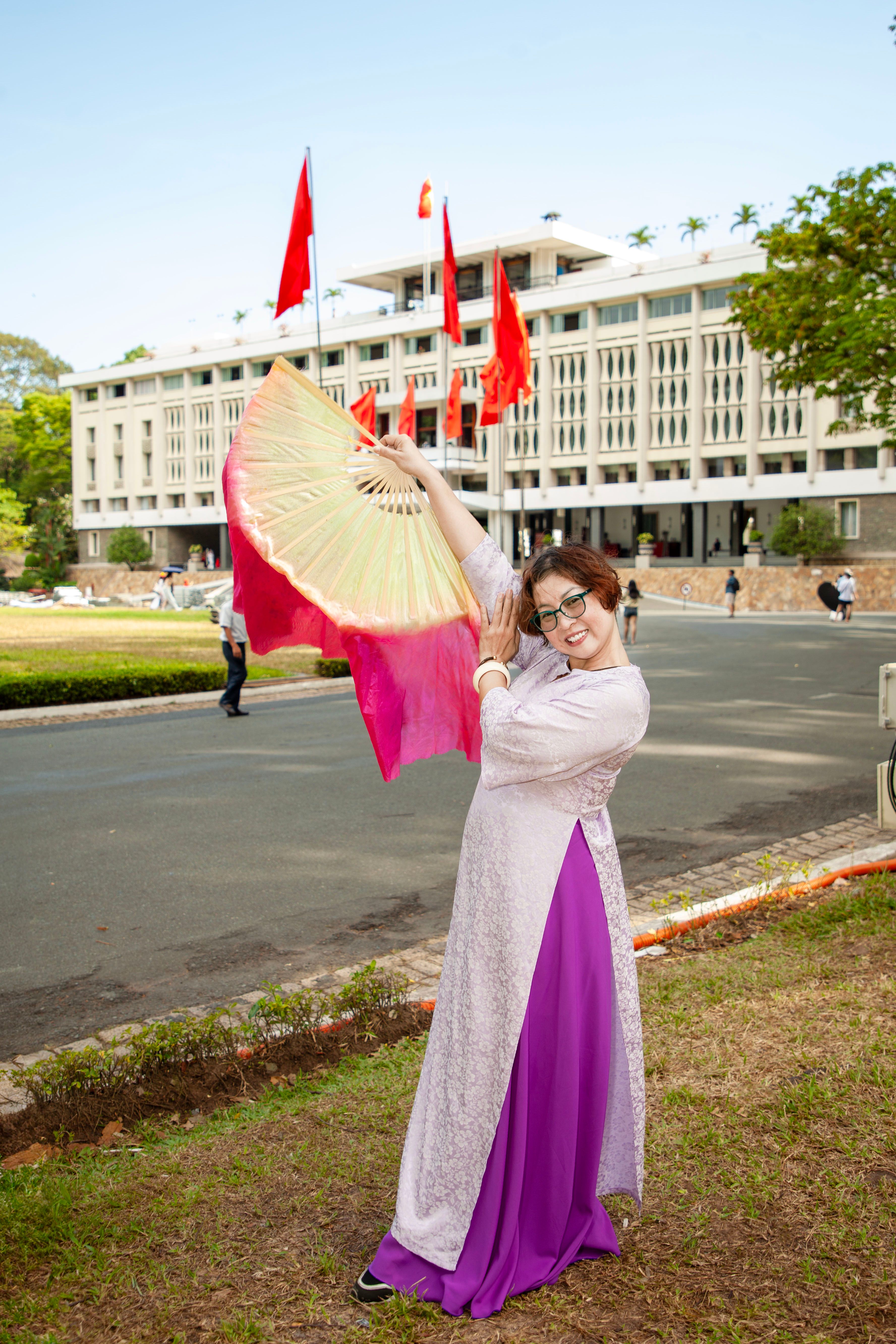 Woman in traditional dress with fan, smiling by landmark building, daytime.