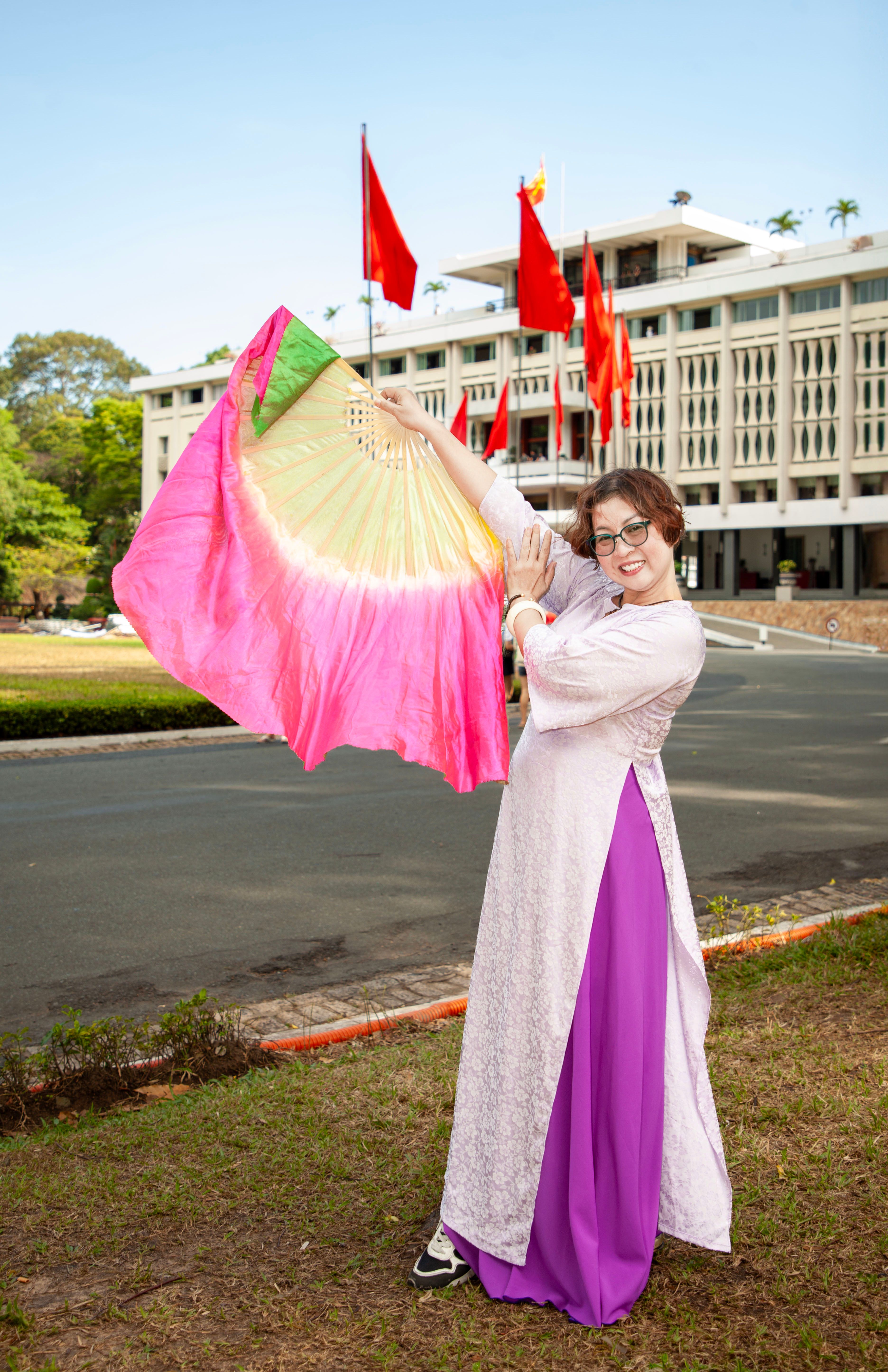 A woman in traditional attire holds a vibrant fan in front of a building with flags.