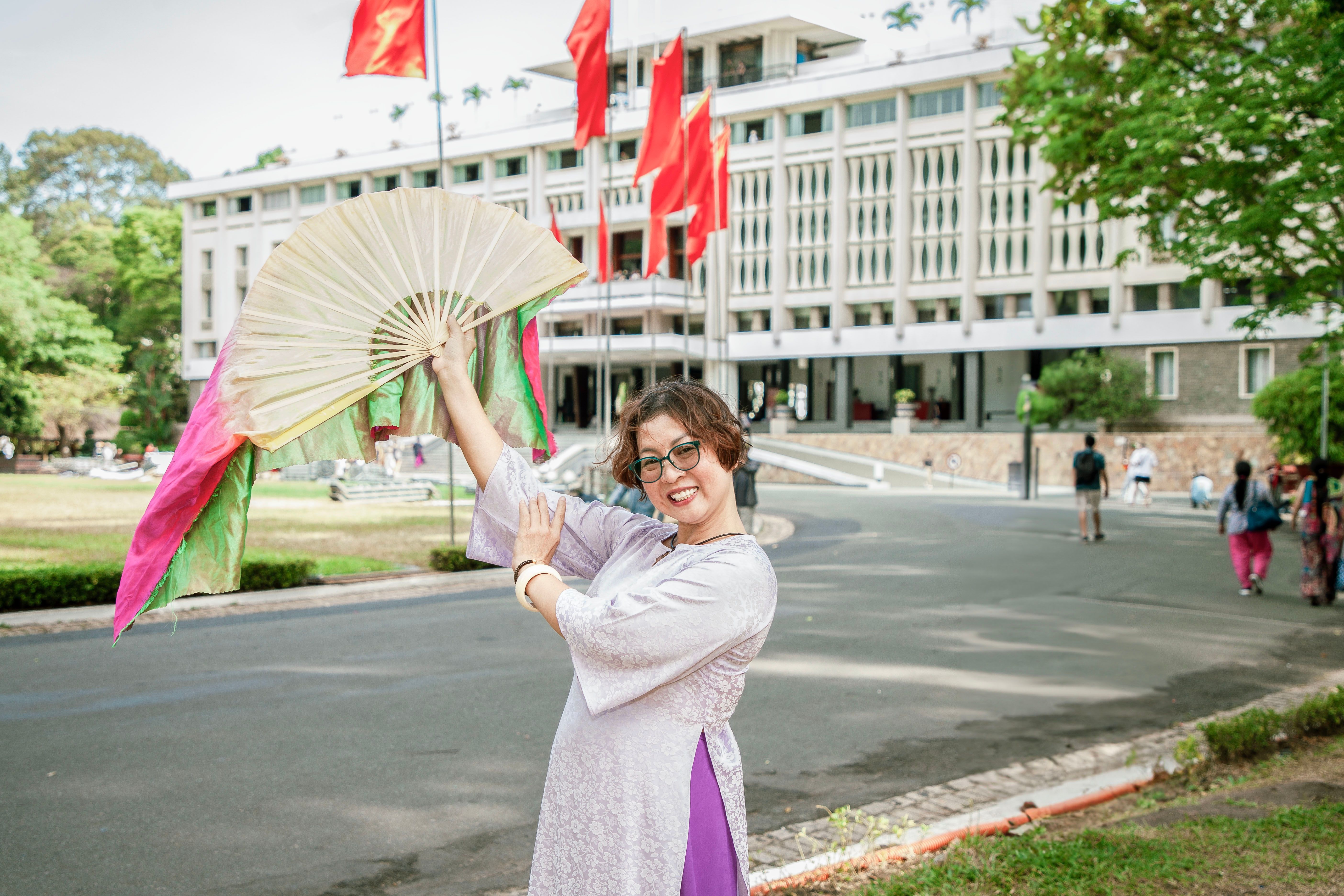 Smiling woman in traditional attire poses with a fan in front of a landmark building with red flags.