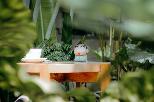 A whimsical toy figure placed on a wooden table surrounded by vibrant greenery in a serene outdoor garden.