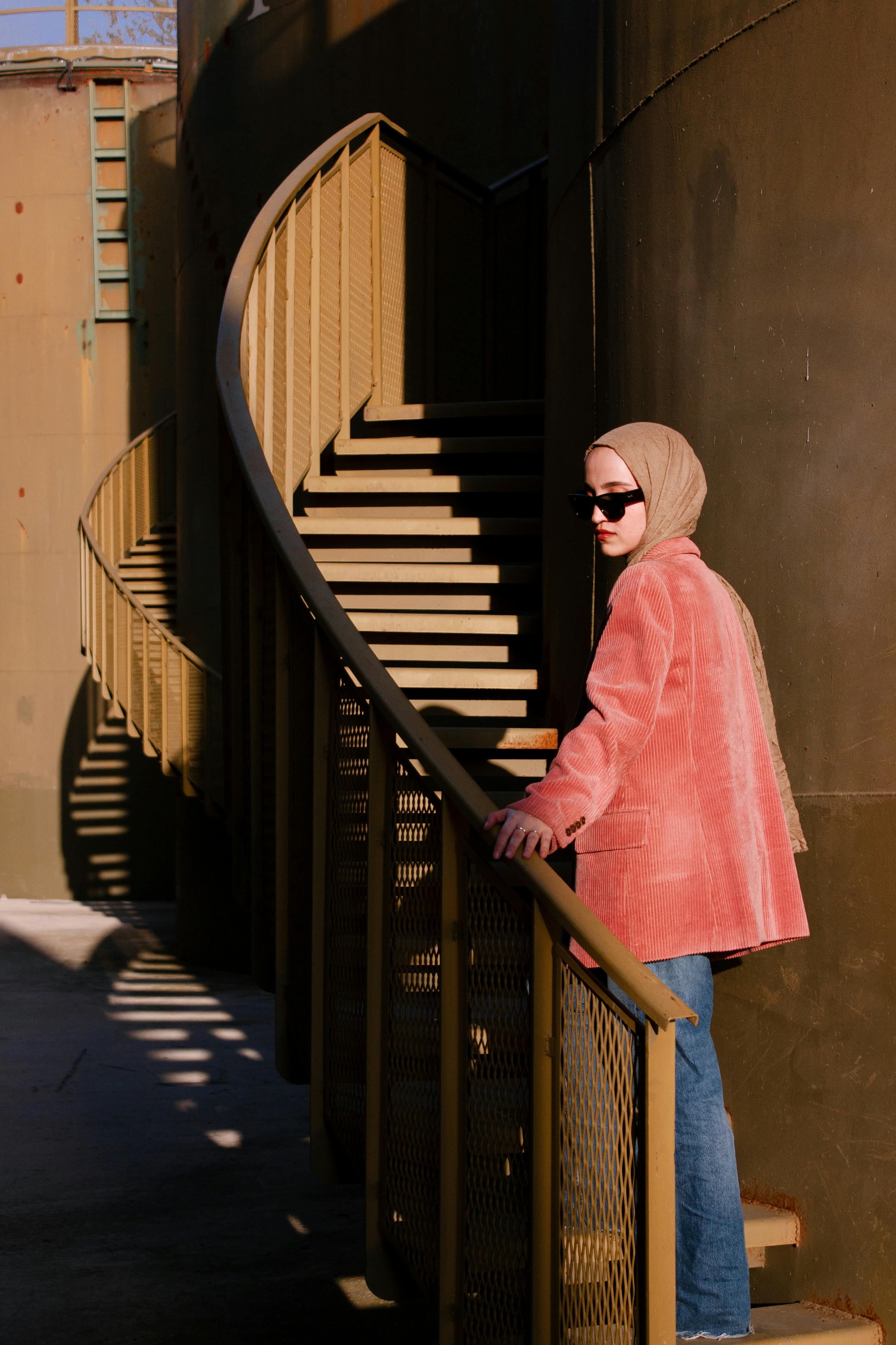 Elegant woman in sunglasses and pink blazer on a modern spiral staircase outside.