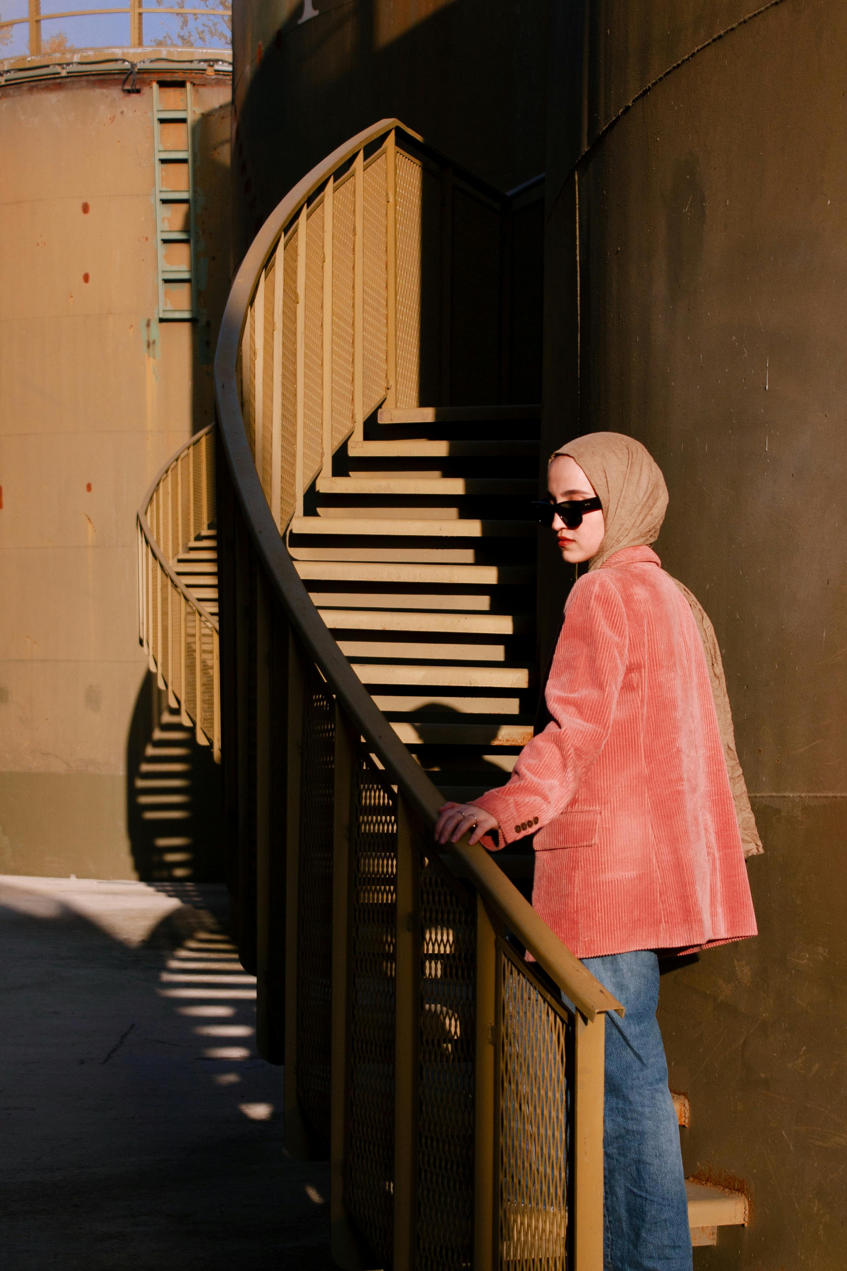 Woman in pink blazer and hijab ascends a modern spiral staircase outdoors.