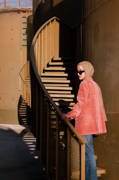 Woman in pink blazer and hijab ascends a modern spiral staircase outdoors.