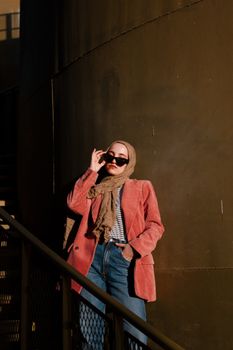 Fashionable woman in hijab wearing sunglasses and a pink jacket, posing outdoors at sunset.