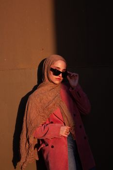 Elegant woman in hijab and sunglasses posing confidently against a textured wall.