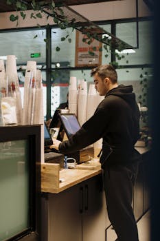 A barista in a black hoodie working at the register in a contemporary café with coffee cups stacked nearby.
