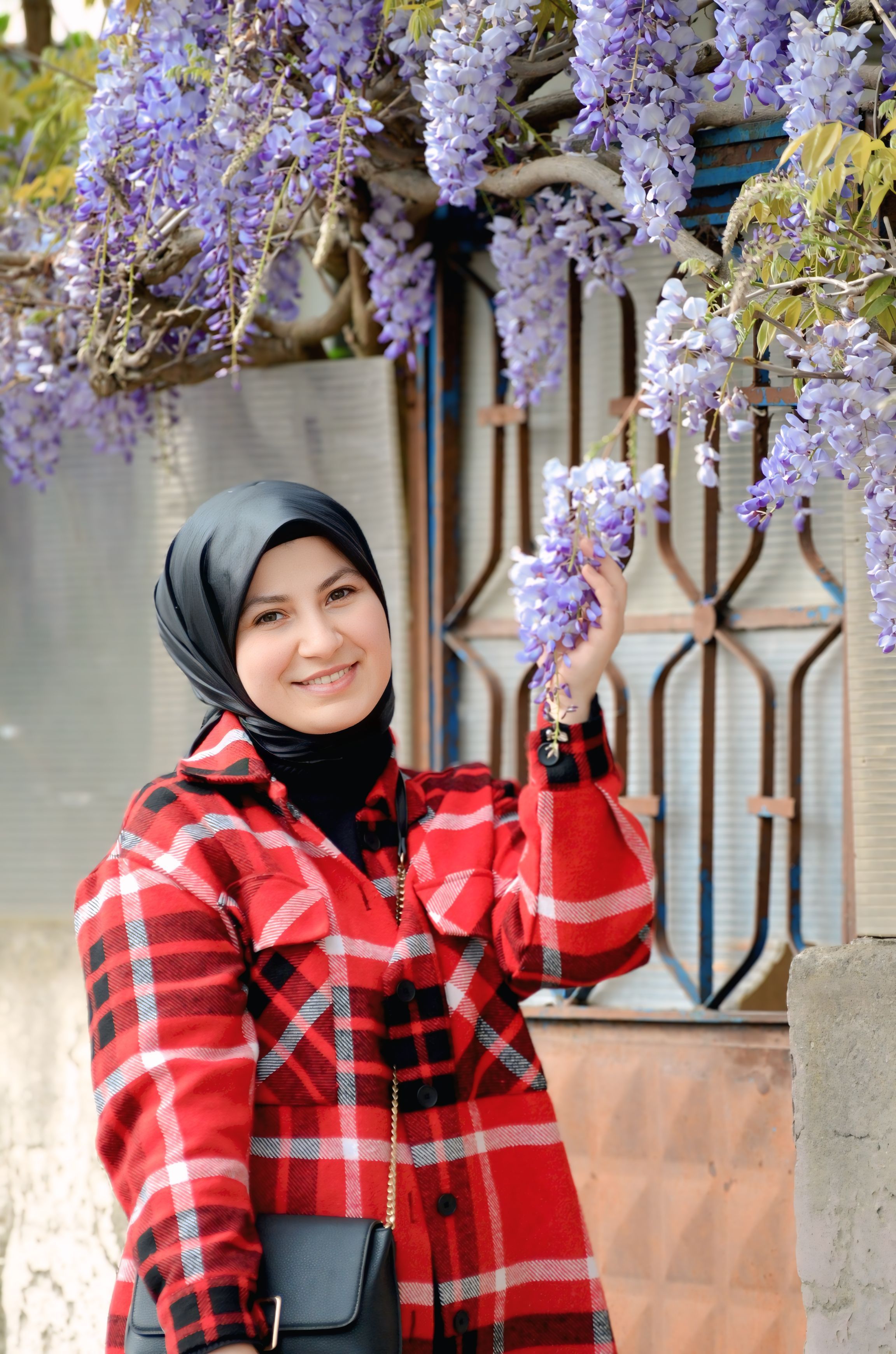 Smiling woman in hijab holding purple wisteria blooms outdoors.