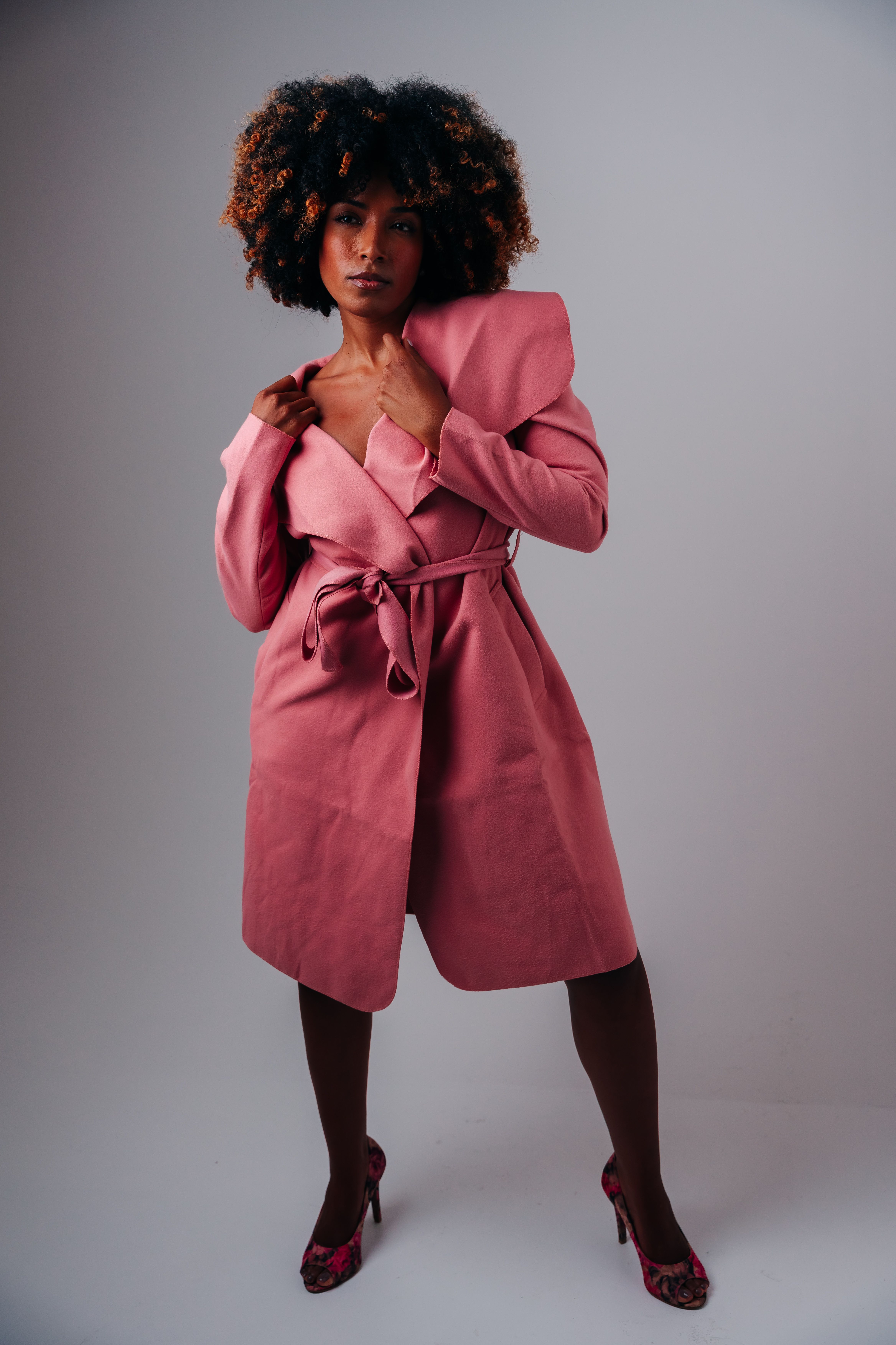 Elegant woman posing in a pink trench coat, full-length studio shot.