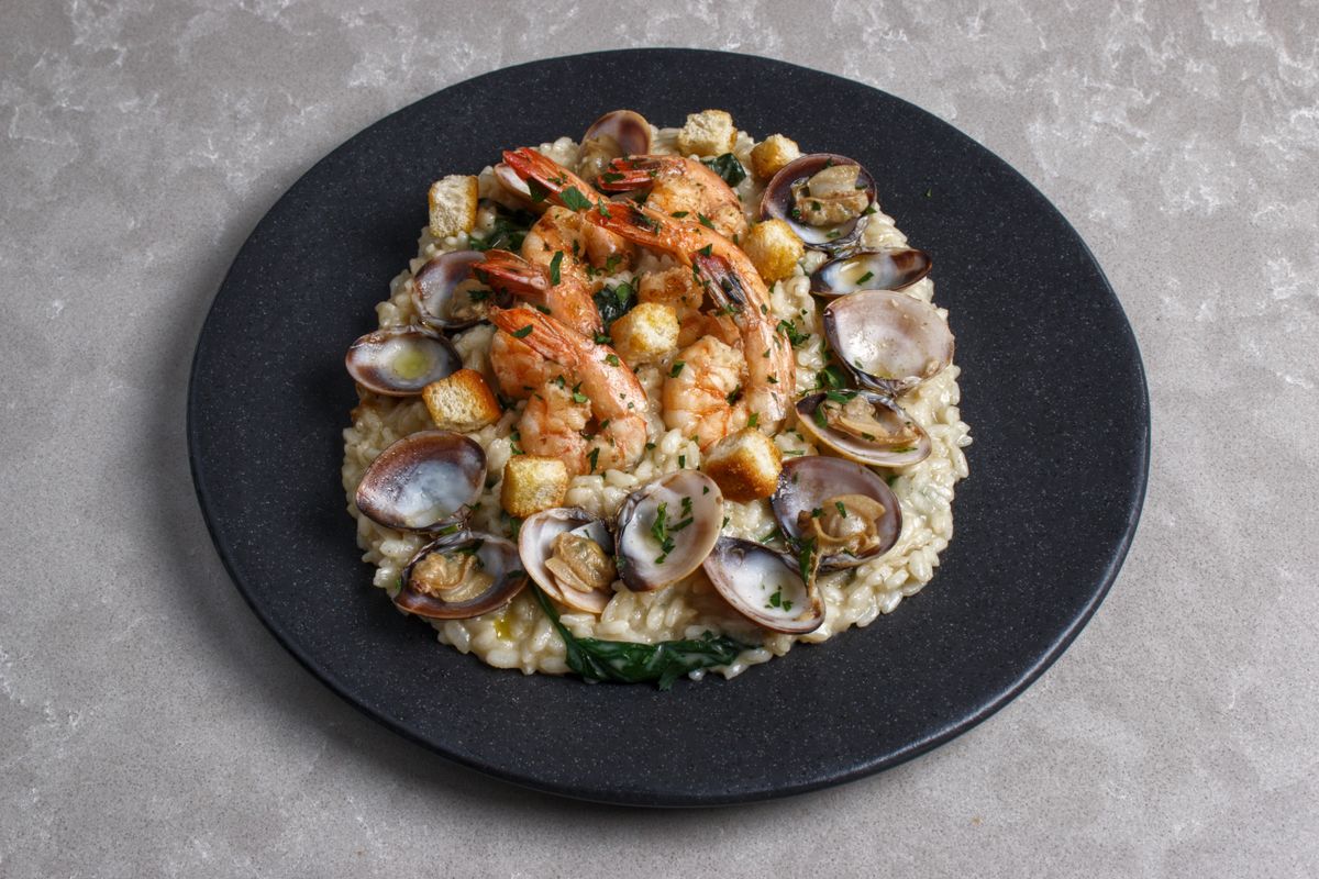 Risotto on a black plate topped with clams in shells, prawns, croutons and greens