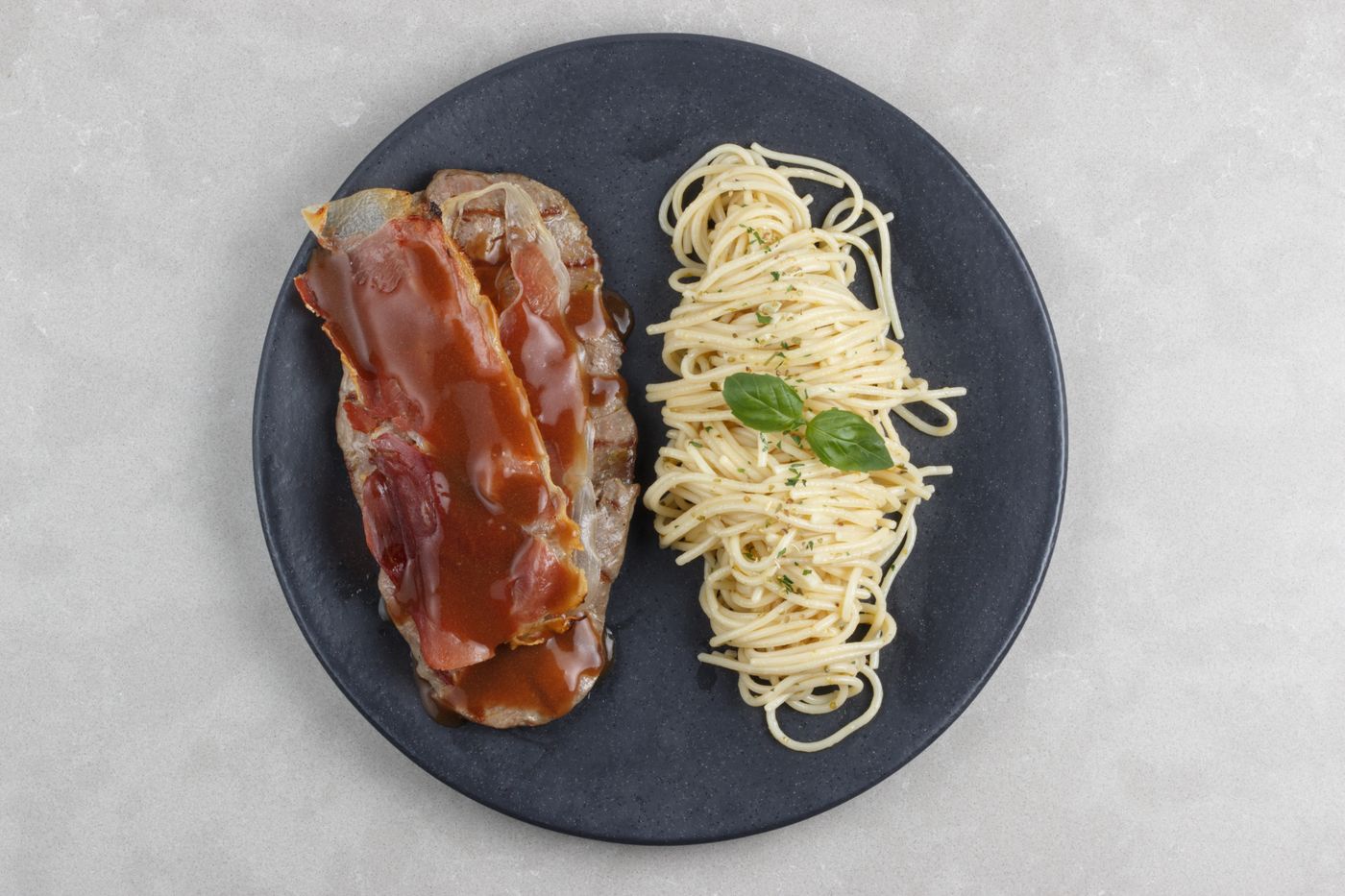 Top view of meatloaf with gravy and spaghetti garnished with basil on a black plate.