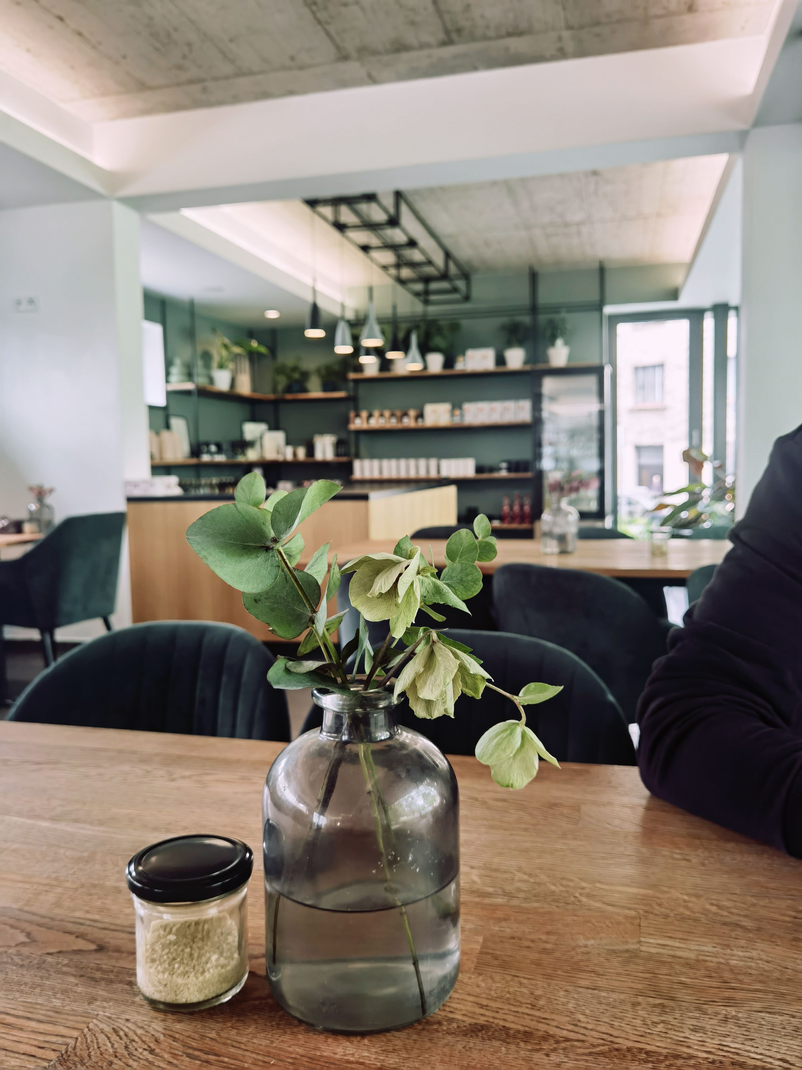 A cozy, modern cafe interior with plants and bookshelves, featuring a vase with leaves on a wooden table.