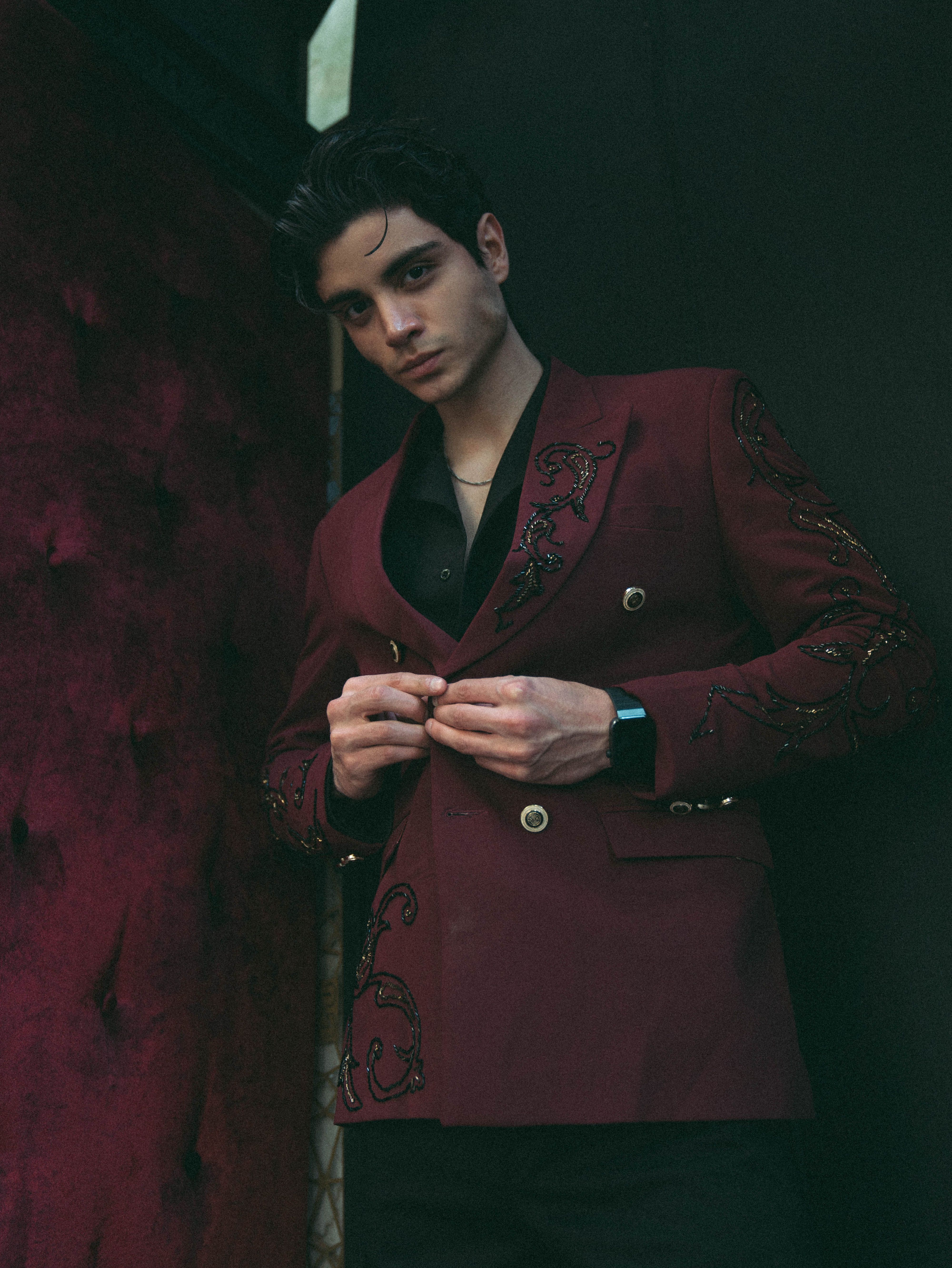 A young man in a red embroidered jacket poses confidently indoors against a dark backdrop.