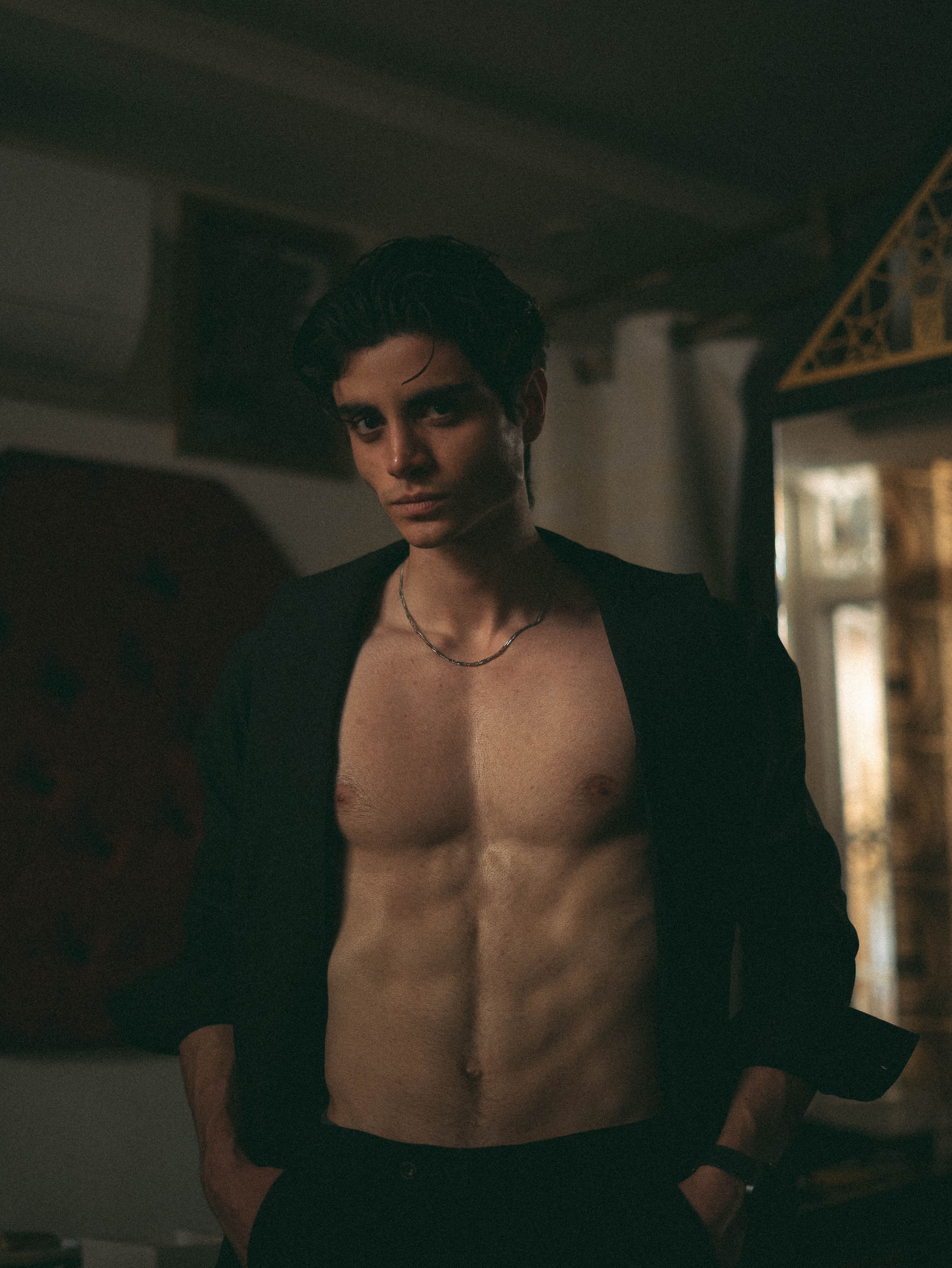 A focused portrait of a young man with defined abs and dark hair in an indoor setting.