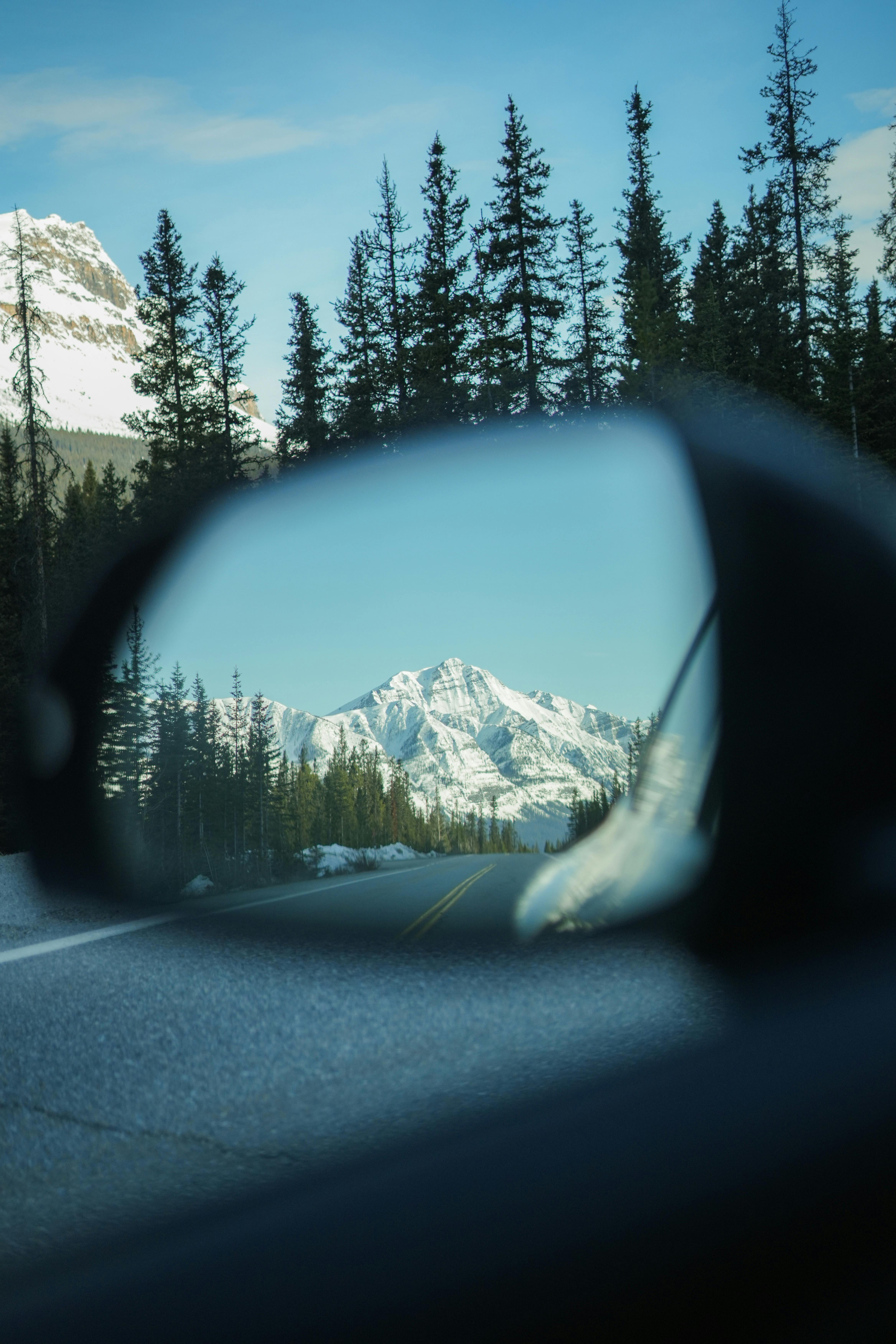 Explore breathtaking snowy mountains as seen through a car mirror on a scenic road trip.