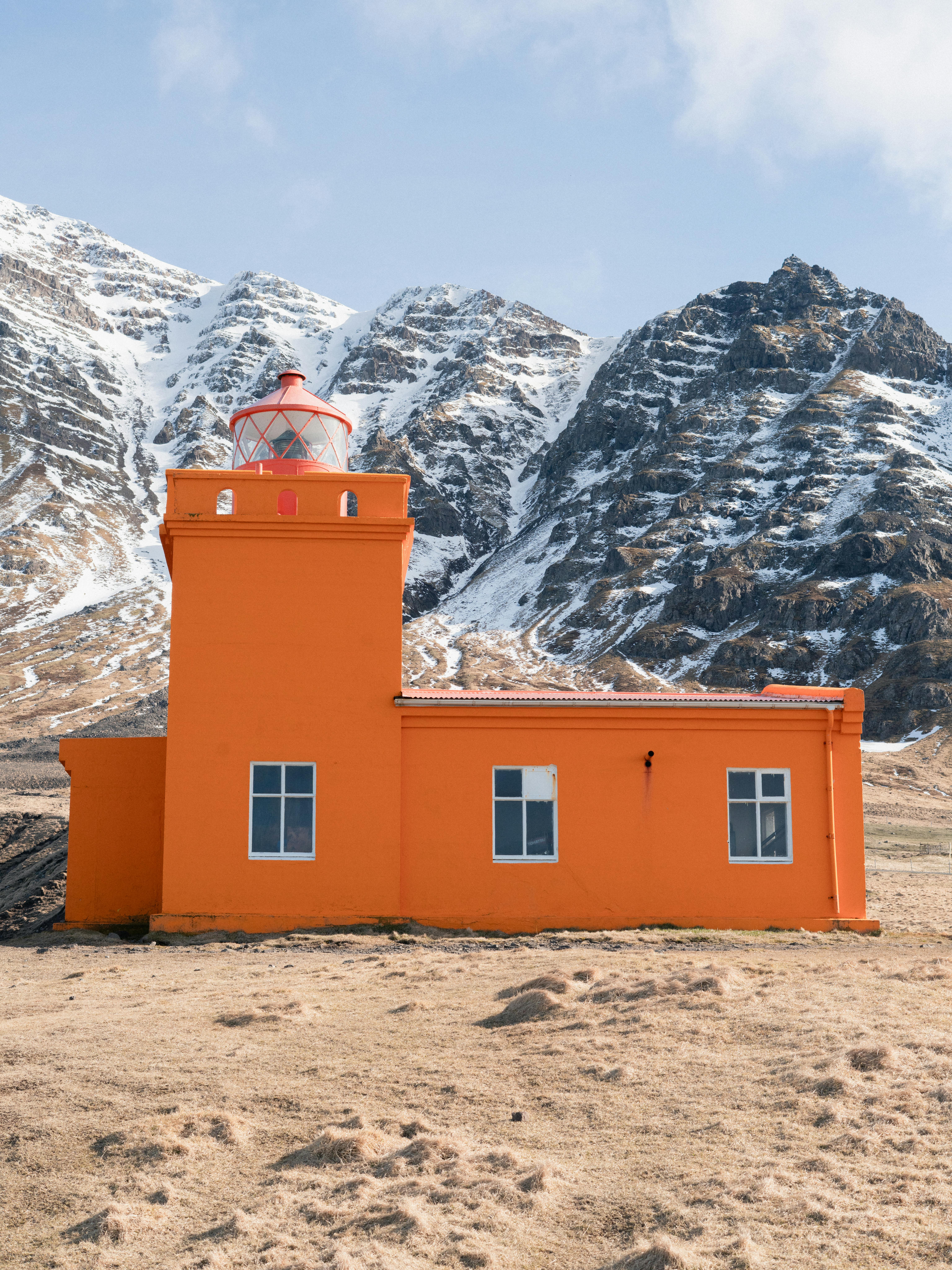 An orange lighthouse stands boldly against snow-capped mountains, creating a stunning contrast.