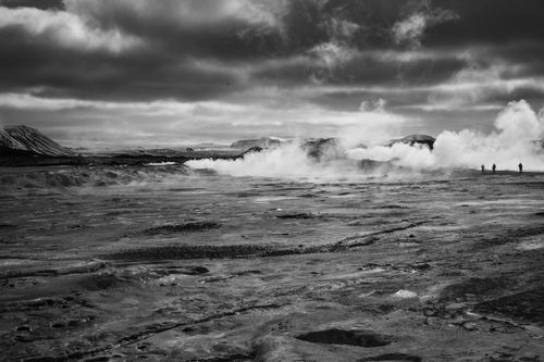 Free Black and white photo of a geothermal landscape in Iceland, capturing steam and distant figures under a moody sky. Stock Photo